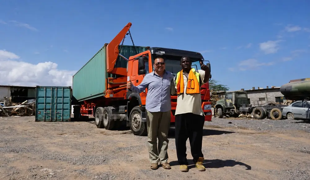 An orange truck-mounted side lifter carrying a green container at an African worksite with two men standing in front.