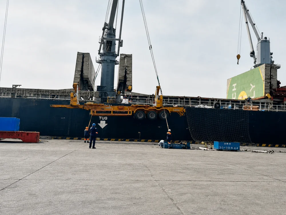 A new side lifter trailer being lowered by a ship's crane onto the quay at a small island port, with dock workers watching.